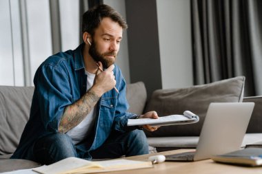 Sakallı Avrupalı adam evdeki kanepede otururken dizüstü bilgisayarla çalışıyor.