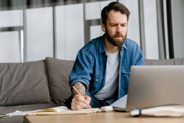 Sakallı adam evde dizüstü bilgisayarla konferans görüşmesi yaparken notlar yazıyor.