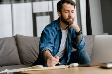 Sakallı adam evde dizüstü bilgisayarla konferans görüşmesi yaparken notlar yazıyor.