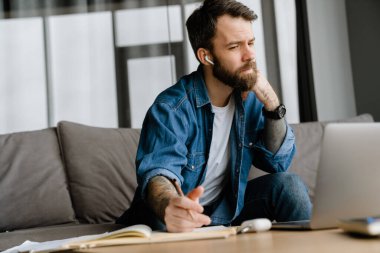 Sakallı Avrupalı adam evdeki kanepede otururken notlar yazıyor.