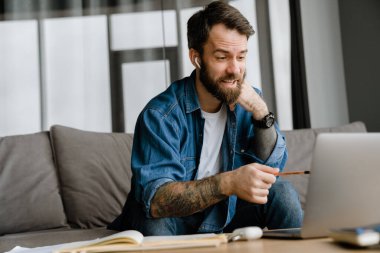 Sakallı Avrupalı adam evdeki kanepede otururken dizüstü bilgisayarla hareket ediyor ve çalışıyor.