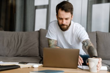 Evde dizüstü bilgisayarla çalışırken kahve içen sakallı Avrupalı adam.