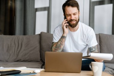 Evde kanepede dizüstü bilgisayarla çalışırken cep telefonuyla konuşan sakallı adam.