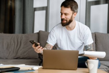 Sakallı Avrupalı adam evdeki kanepede dizüstü bilgisayarla çalışırken cep telefonu kullanıyor.