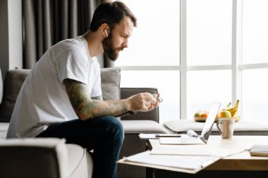 Evde dizüstü bilgisayarla çalışırken kulaklık kullanan Avrupalı sakallı bir adam.