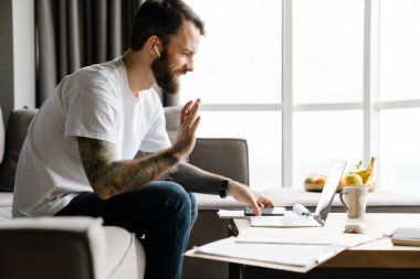 Evde dizüstü bilgisayarla çalışırken kulaklık kullanan sakallı bir adam.