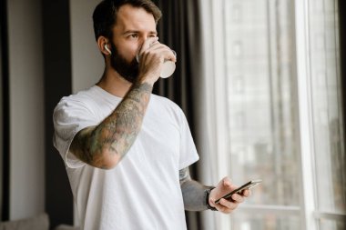 Sakallı Avrupalı adam evde cep telefonu kullanırken kahve içiyor.