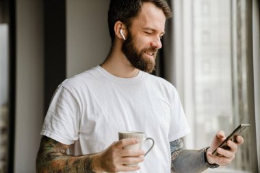 Sakallı Avrupalı adam evde cep telefonu kullanırken kahve içiyor.