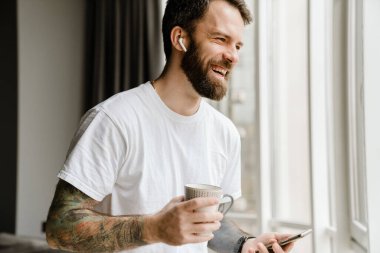 Sakallı Avrupalı adam evde cep telefonu kullanırken kahve içiyor.