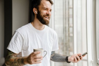 Sakallı Avrupalı adam evde cep telefonu kullanırken kahve içiyor.