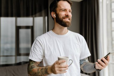 Sakallı Avrupalı adam evde cep telefonu kullanırken kahve içiyor.