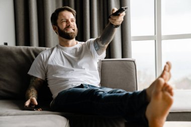 Sakallı Avrupalı adam evde kanepede otururken gülümsüyor ve televizyon seyrediyor.