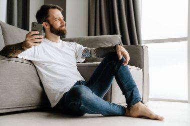 Sakallı Avrupalı adam evde otururken cep telefonu kullanıyor.