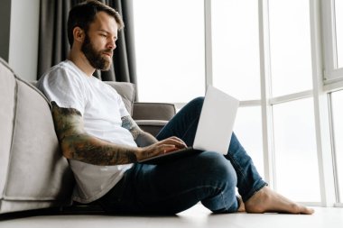 Sakallı Avrupalı adam evde yerde otururken dizüstü bilgisayarla çalışıyor.