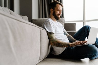 Sakallı Avrupalı adam evde yerde otururken dizüstü bilgisayarla çalışıyor.