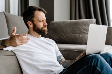 Sakallı Avrupalı adam evde yerde otururken dizüstü bilgisayarla çalışıyor.