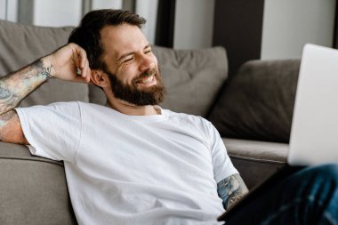 Sakallı Avrupalı adam evde yerde otururken dizüstü bilgisayarla çalışıyor.