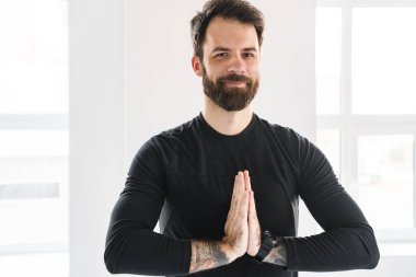 Gülümseyen orta yaşlı beyaz sporcu. Parlak bir odada egzersiz yapıyor. Avuçlar birbirine girmiş meditasyon yapıyor.