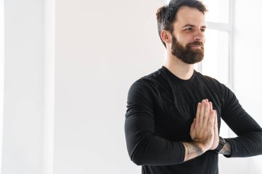 Gülümseyen orta yaşlı beyaz sporcu. Parlak bir odada egzersiz yapıyor. Avuçlar birbirine girmiş meditasyon yapıyor.