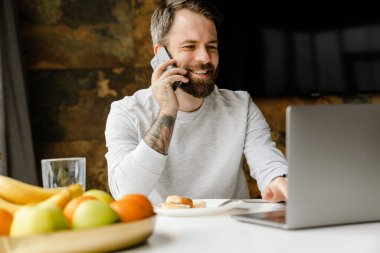 Sakallı adam içeride dizüstü bilgisayarla çalışırken gülümsüyor ve telefonla konuşuyor.