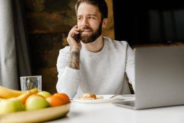 Evde dizüstü bilgisayarla çalışırken cep telefonuyla konuşan sakallı bir adam.