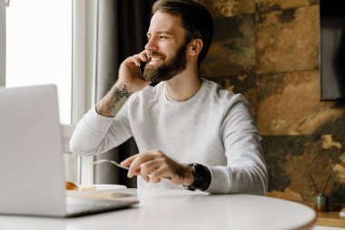 Sakallı adam içeride dizüstü bilgisayarla çalışırken gülümsüyor ve telefonla konuşuyor.