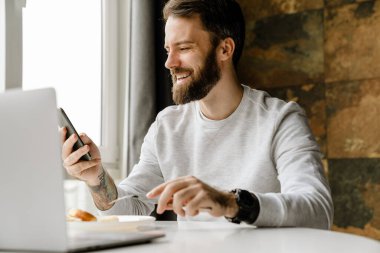 Sakallı adam içeride dizüstü bilgisayarla çalışırken gülümsüyor ve cep telefonu kullanıyor.