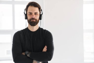 Genç, sırıtan esmer, sakallı, spor kıyafetli beyaz adam içeride durmuş kulaklıkla müzik dinliyor.