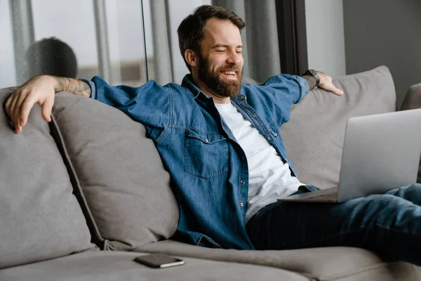 Sakallı Avrupalı adam evdeki kanepede otururken dizüstü bilgisayarla çalışıyor.