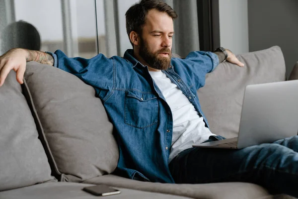 Sakallı Avrupalı adam evdeki kanepede otururken dizüstü bilgisayarla çalışıyor.