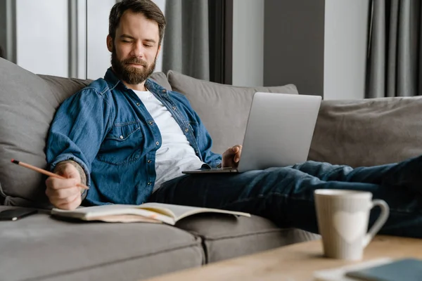 Sakallı adam evdeki kanepede dizüstü bilgisayarla çalışırken notlar yazıyor.