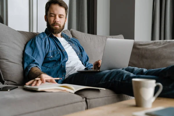 Bärtiger Mann Schreibt Notizen Während Hause Mit Laptop Auf Dem — Stockfoto