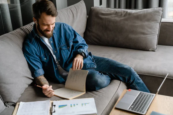 Sakallı Avrupalı adam evdeki kanepede otururken notlar yazıyor.