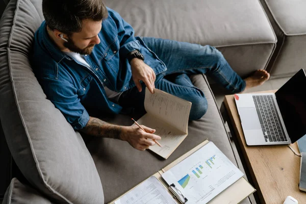 Sakallı adam evdeki kanepede dizüstü bilgisayarla çalışırken notlar yazıyor.