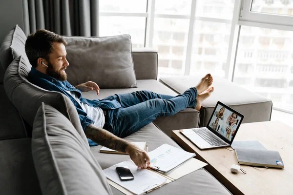 Sakallı adam evdeki kanepede otururken bilgisayarla konferans görüşmesi yapıyor.