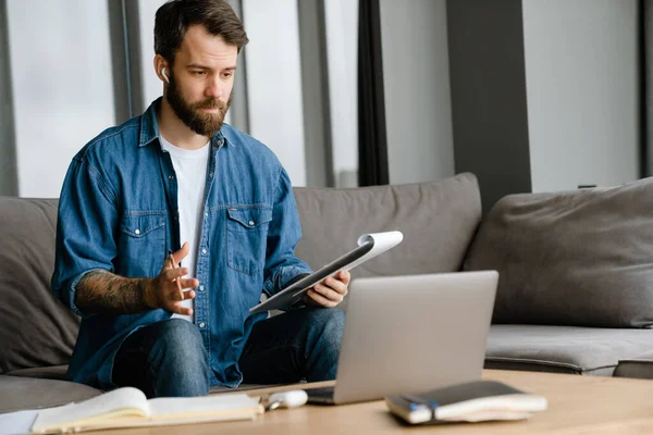 Sakallı Avrupalı adam evdeki kanepede otururken dizüstü bilgisayarla çalışıyor.