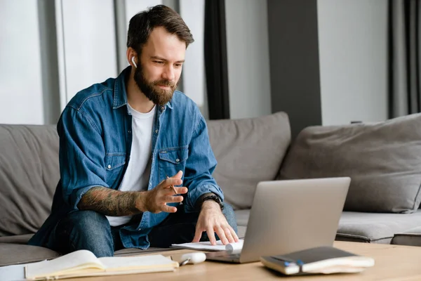 Sakallı Avrupalı adam evdeki kanepede otururken dizüstü bilgisayarla çalışıyor.