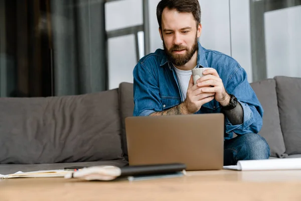 Bärtiger Europäischer Mann Trinkt Kaffee Während Hause Mit Laptop Arbeitet — Stockfoto