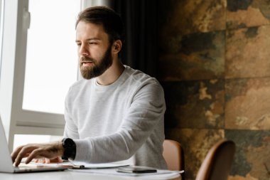 Sakallı adam içeride masada otururken bilgisayar ve kağıtlarla çalışıyor.