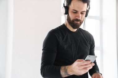 Genç, esmer, sakallı, spor kıyafetli beyaz bir adam. Evde durmuş, kulaklıkla, elinde cep telefonuyla müzik dinliyor.