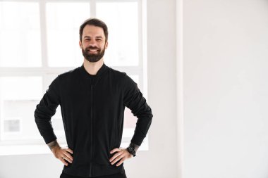 Gülümseyen esmer, beyaz bir adam spor giyim kuşamında evde dikiliyor.