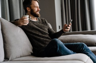 Sakallı adam evde kanepede otururken cep telefonu kullanıyor ve kahve içiyor.