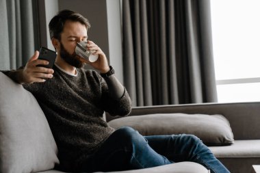 Sakallı adam evde kanepede otururken cep telefonu kullanıyor ve kahve içiyor.