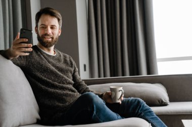 Sakallı adam evde kanepede otururken cep telefonu kullanıyor ve kahve içiyor.