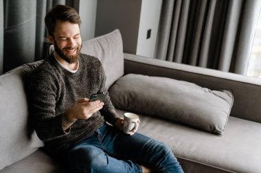 Sakallı adam evde kanepede otururken cep telefonu kullanıyor ve kahve içiyor.