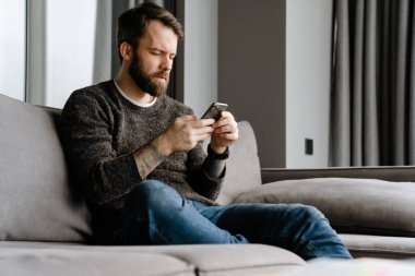 Sakallı Avrupalı adam evdeki koltukta otururken cep telefonu kullanıyor.