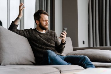 Sakallı Avrupalı adam evdeki koltukta otururken cep telefonu kullanıyor.