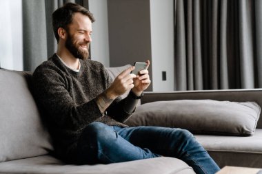 Sakallı Avrupalı adam evdeki koltukta otururken cep telefonu kullanıyor.