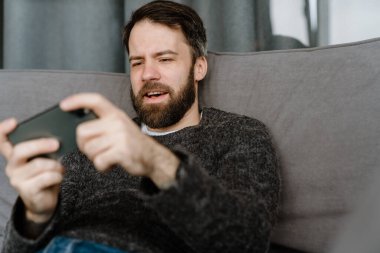 Sakallı Avrupalı adam evdeki koltukta otururken cep telefonu kullanıyor.