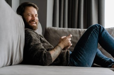 Sakallı Avrupalı adam evdeki koltukta otururken cep telefonu kullanıyor.